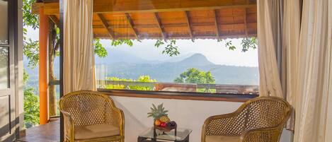 A room with two wicker chairs, a small table with fruit, and a large window offering a mountain view.