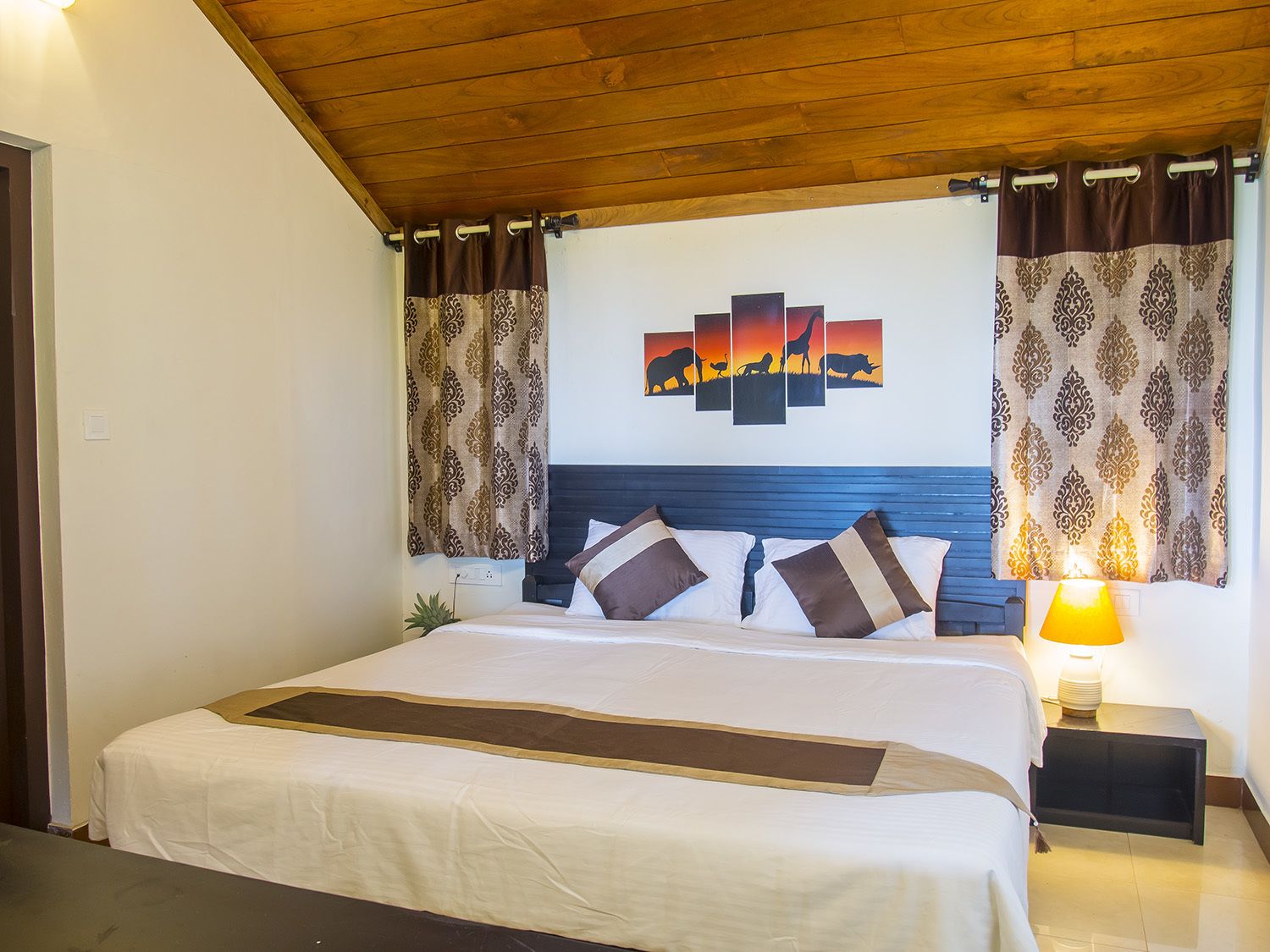 A neatly made bed with a headboard, a bedside lamp, and a wall art depicting a sunset with silhouettes of animals.