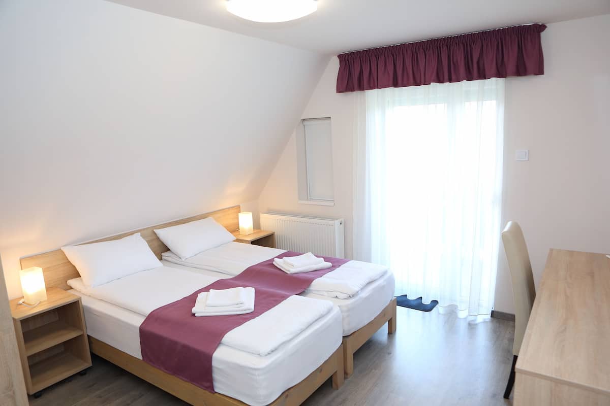Uma cama de casal com lençóis brancos e um cobre-leito bordô, duas mesas de cabeceira com luminárias, uma escrivaninha de madeira e uma janela com cortinas translúcidas.