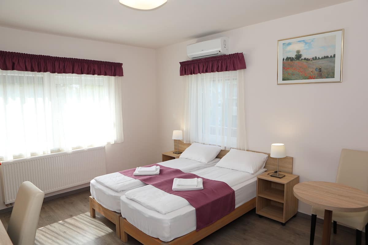Uma cama de casal com roupa de cama branca e detalhes em roxo, duas mesas de cabeceira com abajures, uma pequena mesa de madeira e um quadro na parede.