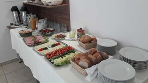 Una mesa de buffet con diversos alimentos, como carnes cortadas, verduras y panes, junto con varios dispensadores de bebidas y platos apilados.