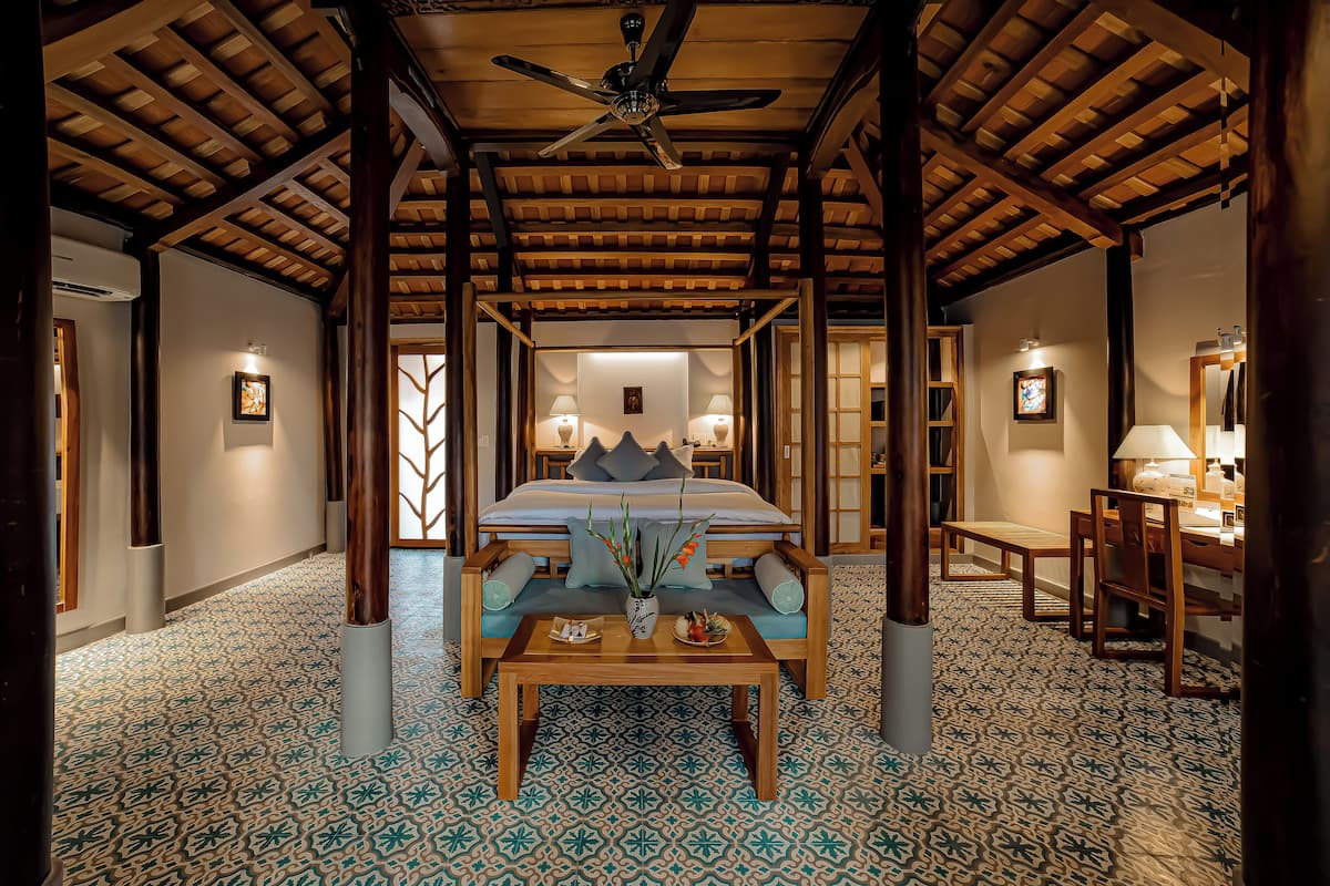 A spacious room with a large bed, a wooden ceiling fan, and a patterned floor.