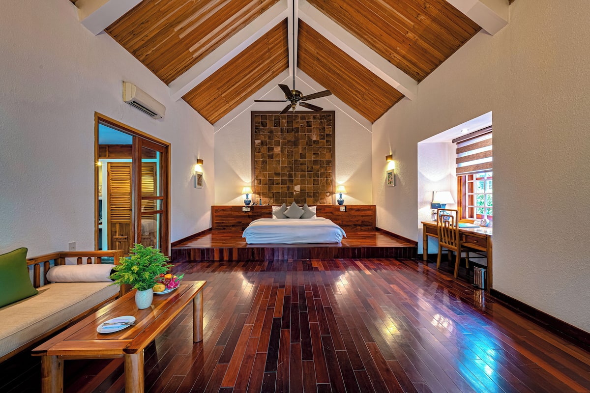 A spacious bedroom with a large bed, wooden ceiling beams, a dining area, and a view of the outdoors.