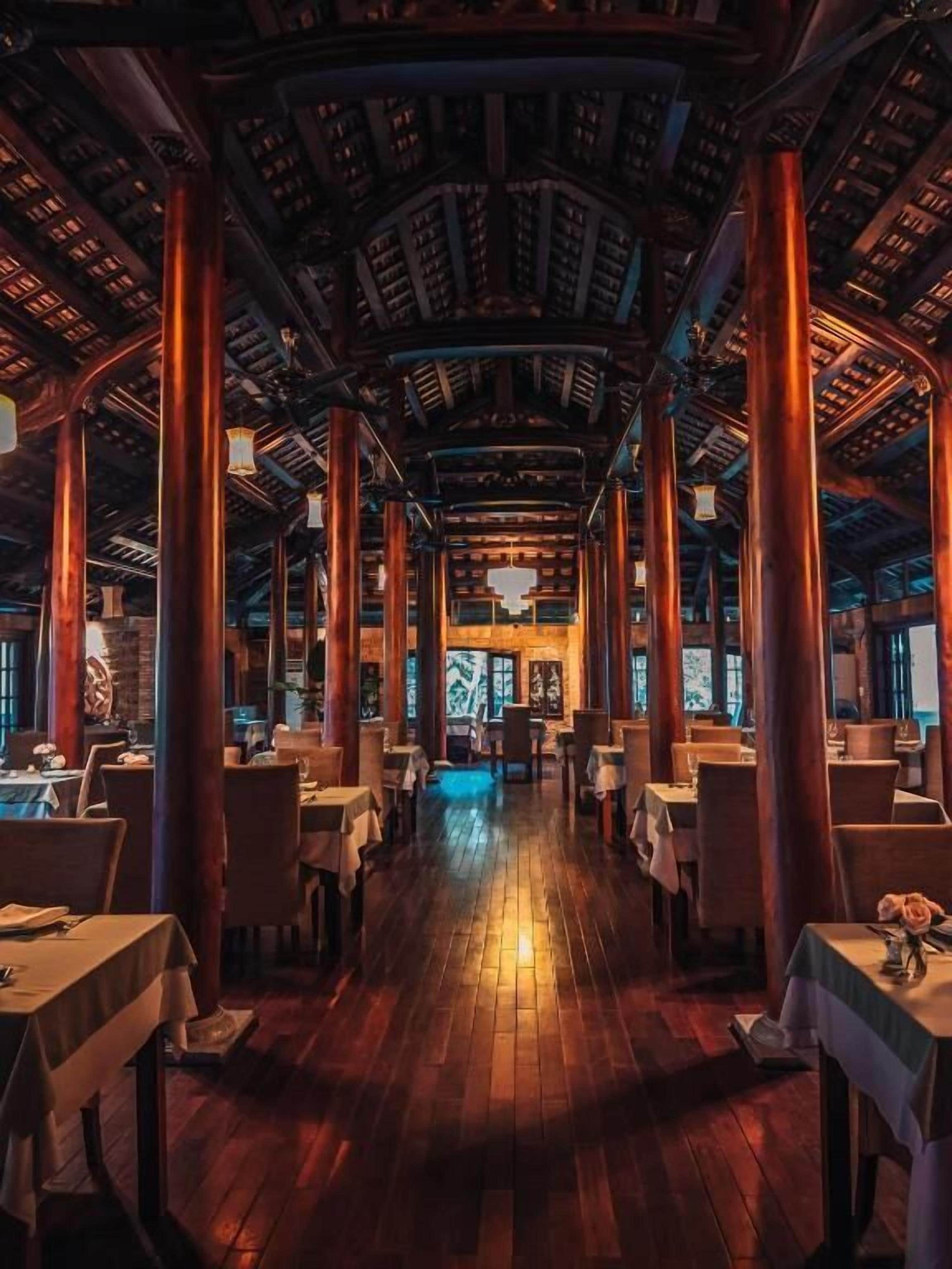 A restaurant with a central open space, wooden pillars, and tables arranged on either side.