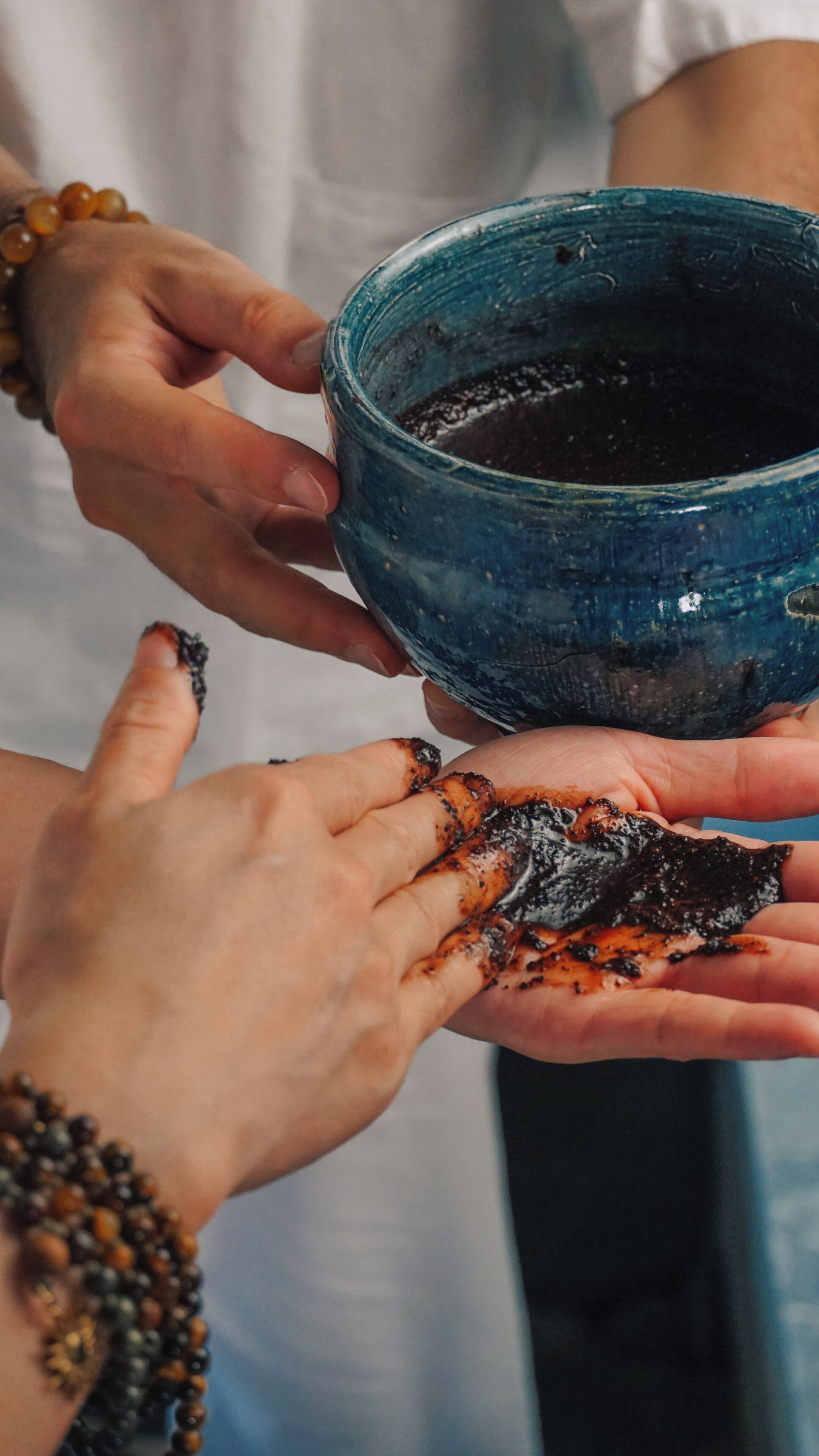A person holding a blue bowl with a dark substance, another person with a hand covered in the substance, and a third person with a bracelet.