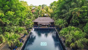 A rectangular swimming pool with two blue square markings, surrounded by lush greenery and palm trees, with a small building in the background. - Pilgrimage Wellness Retreat – part of Pilgrimage Village Resort & Spa (Hue)
