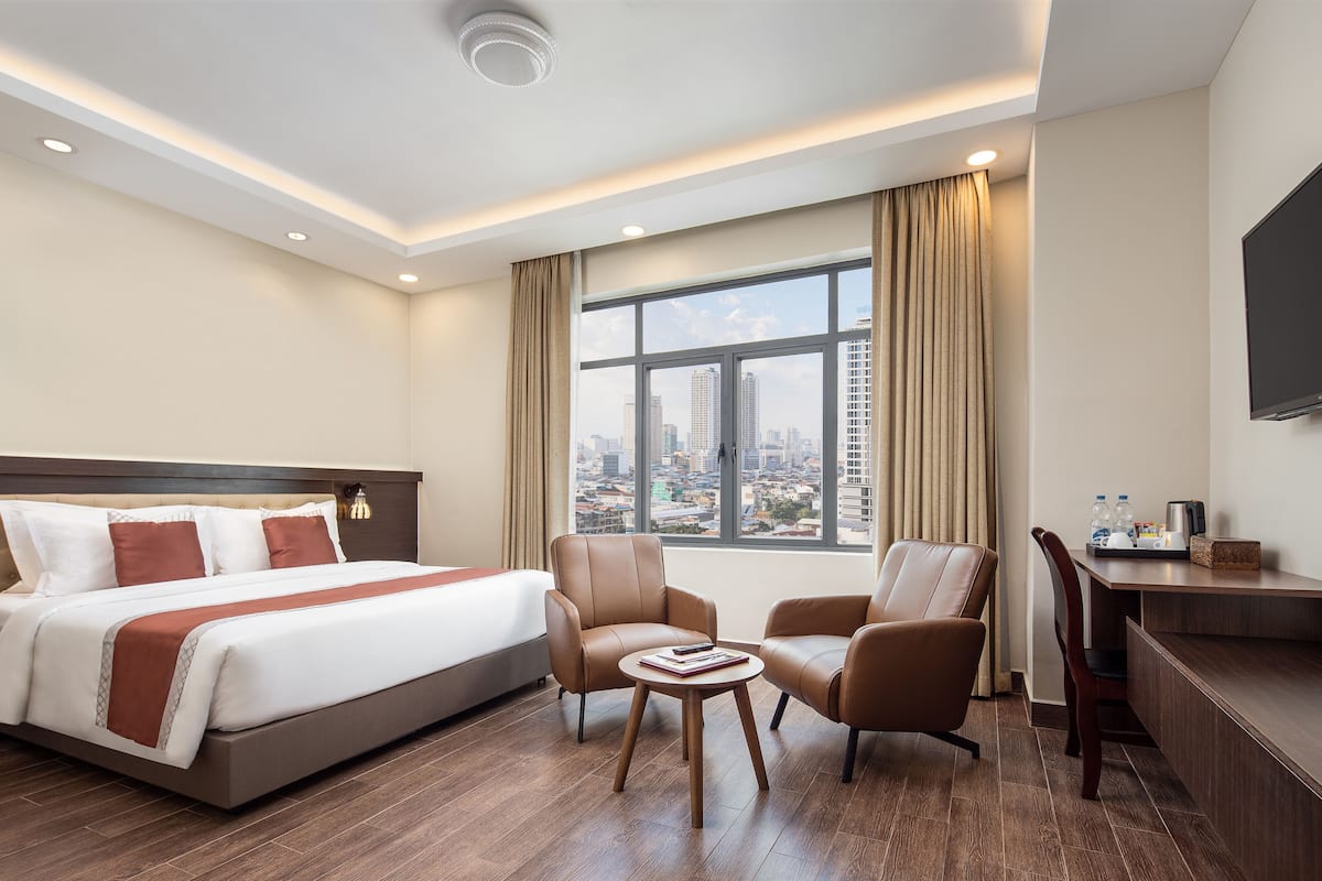 Une chambre d’hôtel avec un grand lit, deux fauteuils, une petite table, un bureau et une grande fenêtre donnant sur la ville.