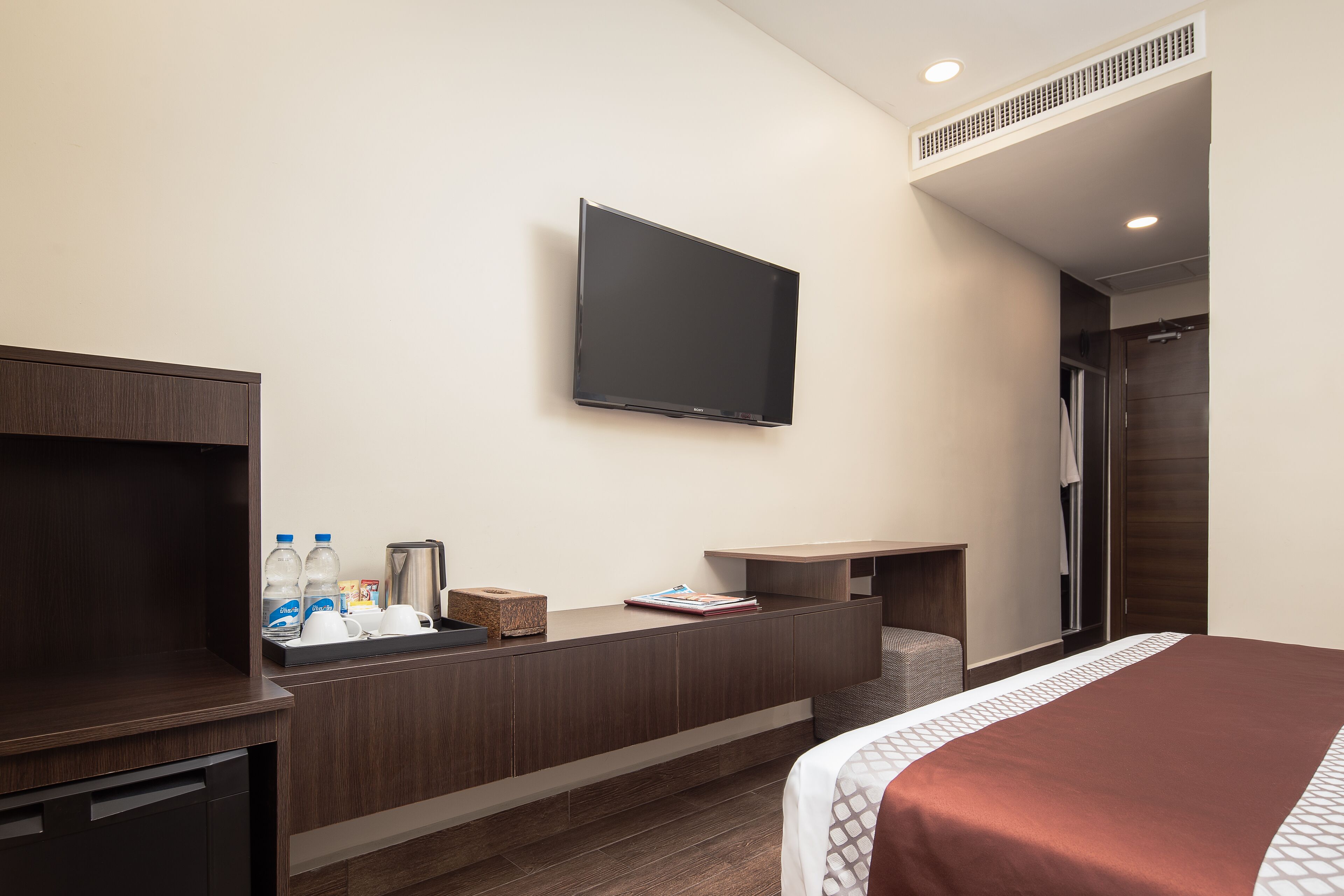 A hotel room with a bed, a TV mounted on the wall, a desk with a coffee maker, and a small seating area.