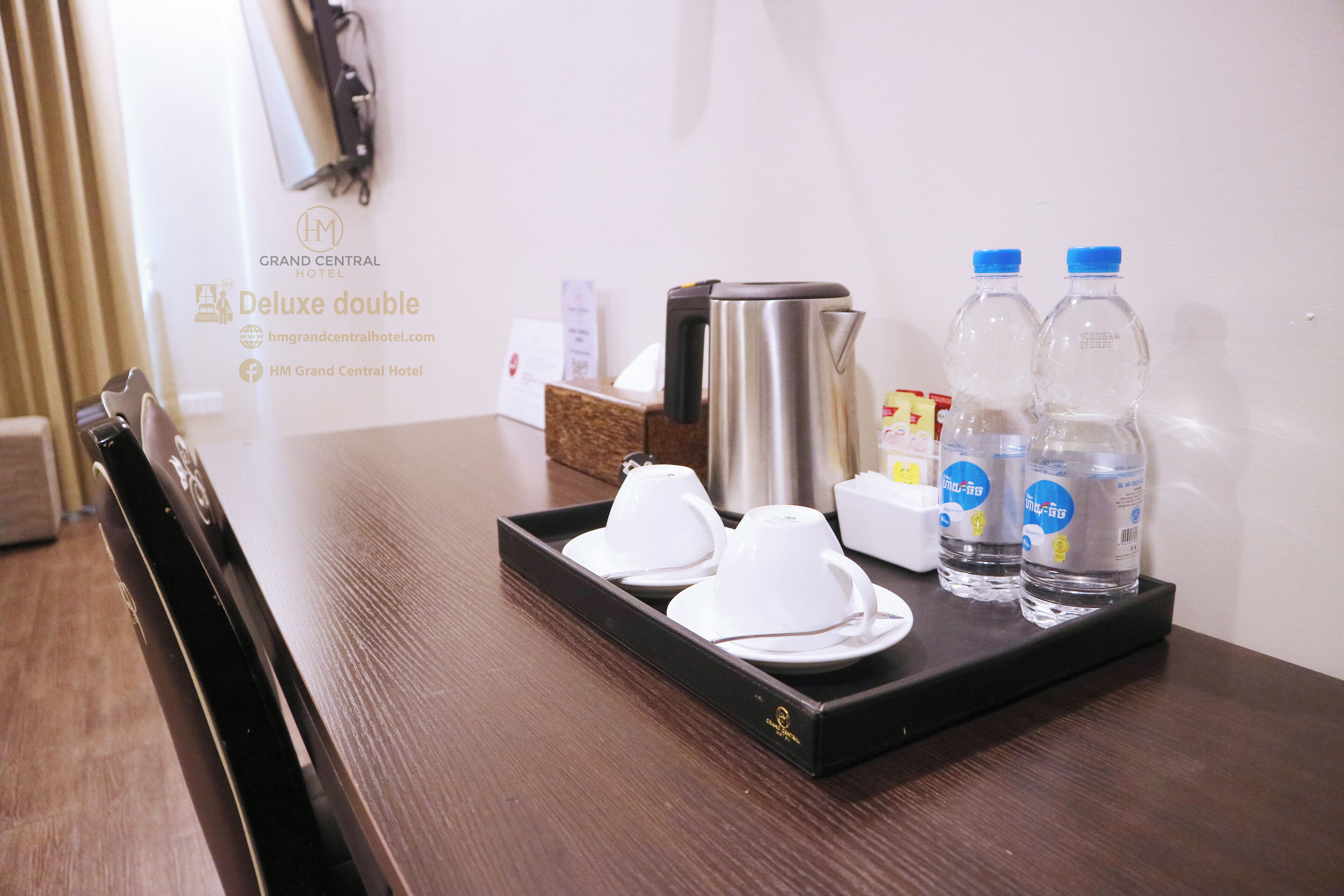 A hotel room with a wooden table set with white cups and saucers, two water bottles, and a coffee pot.