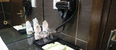 A hotel bathroom with a hairdryer mounted on the wall, a tray with toiletries, and a mirror.
