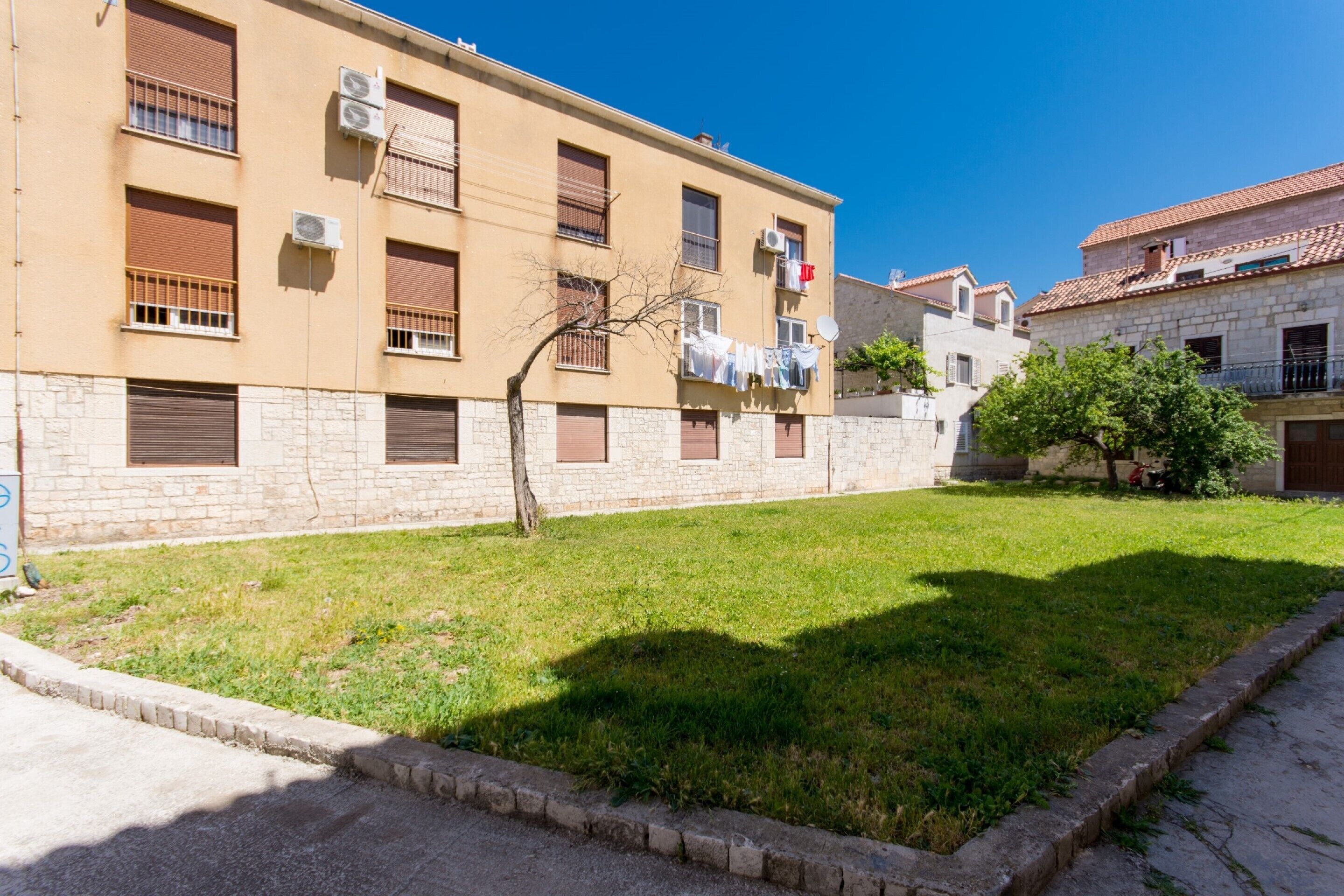 A residential building with multiple balconies, air conditioning units, and a small grassy area in front.