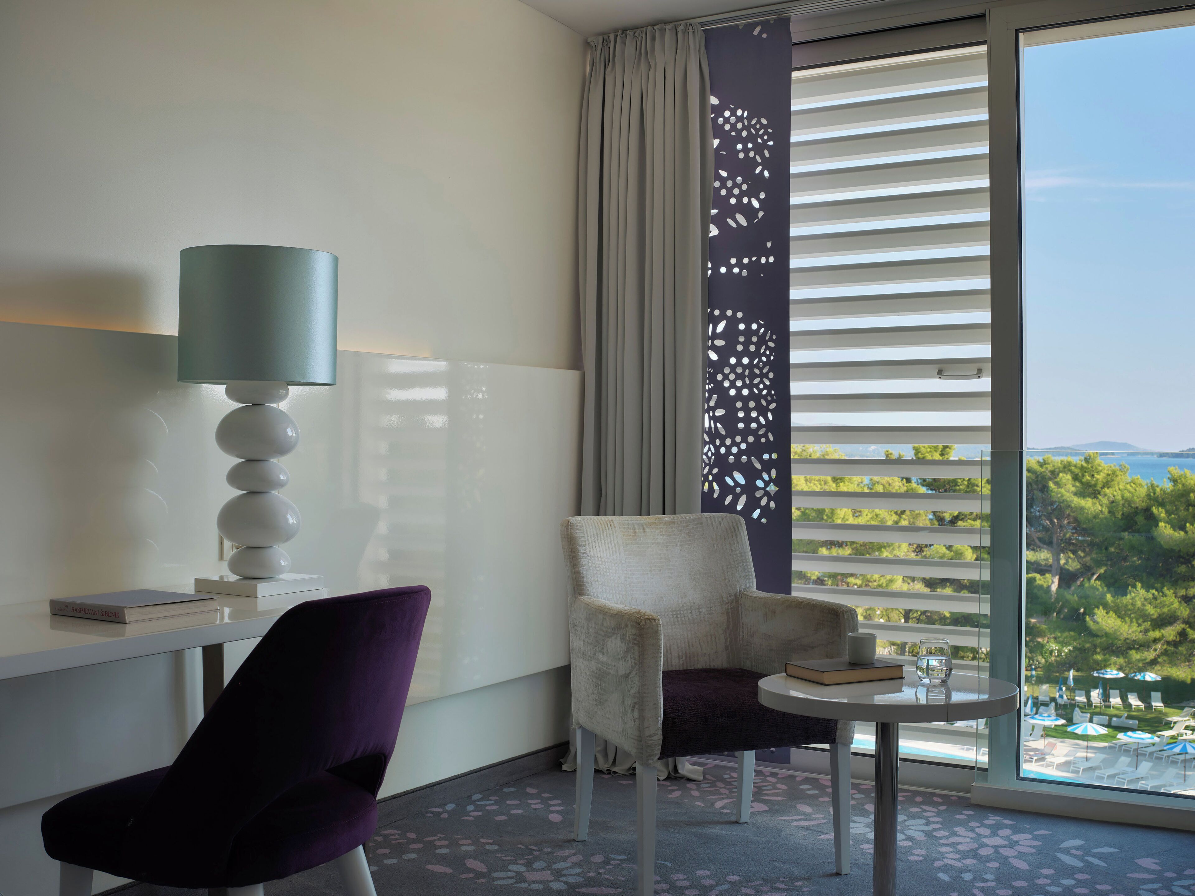 Habitación de hotel con escritorio, una silla, una lámpara, una mesita y vista a una piscina y follaje.