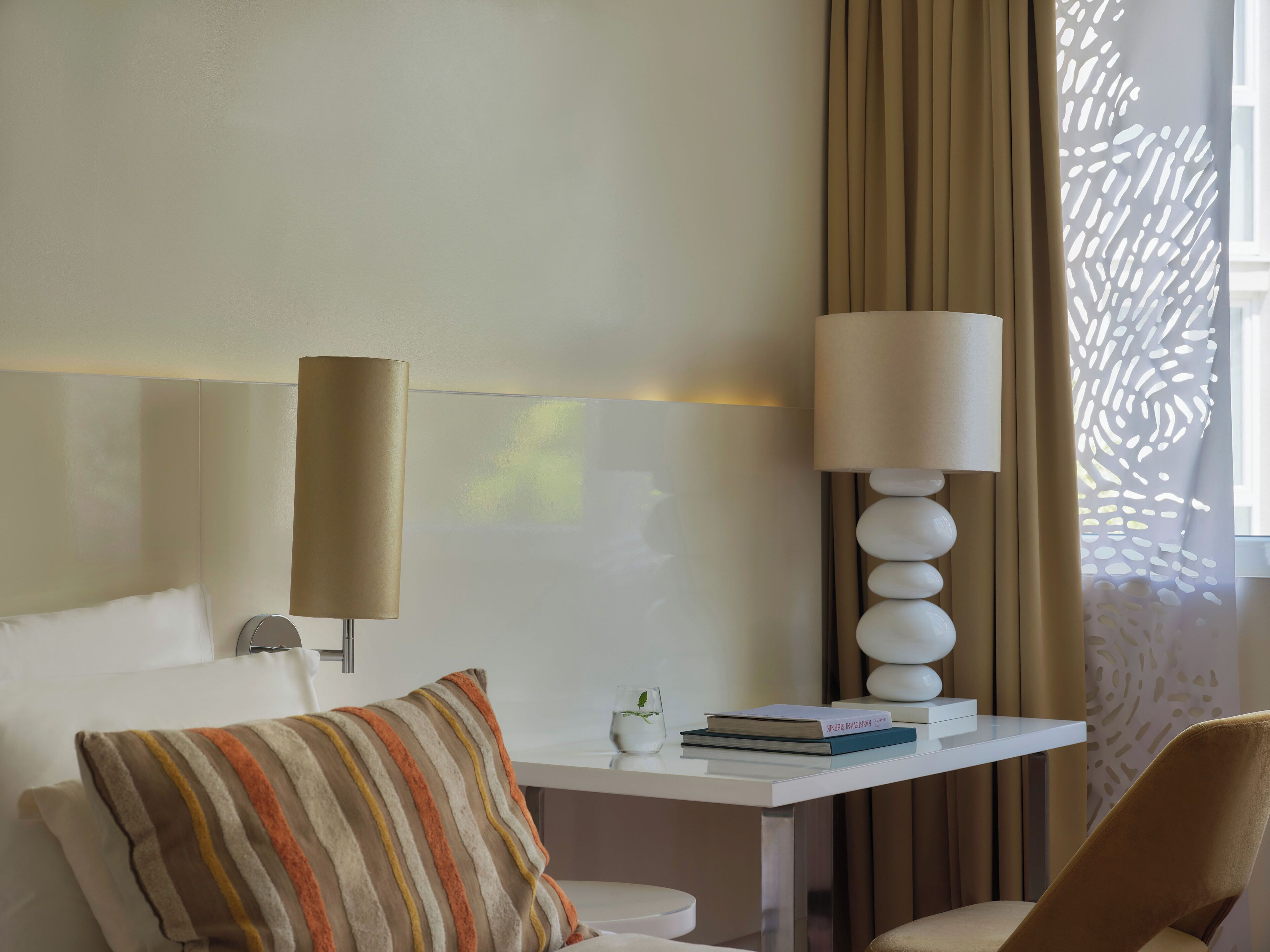 Ein modernes Hotelzimmer mit einem weißen Sofa, einem gestreiften Kissen, einem Beistelltisch mit Büchern und einer Lampe sowie einem dekorativen Blick aus dem Fenster.
