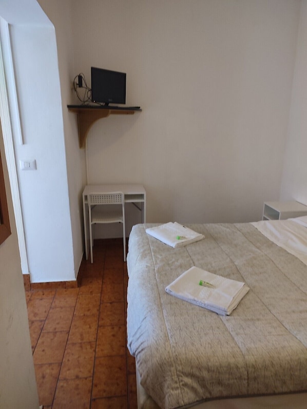 Una habitación de hotel con cama, un escritorio con ordenador y una pequeña estantería encima.