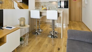 Un espacio habitable compacto con televisión, una pequeña zona de bar con taburetes, un sofá gris y una cocina al fondo.