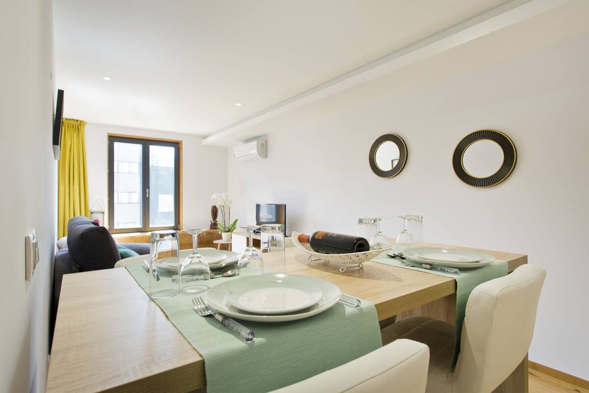 A dining area with a wooden table set for four, a television in the background, and a window with yellow curtains.