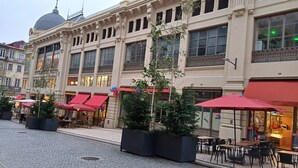 Un edificio histórico con amplios ventanales y una cúpula, que cuenta con zonas de estar al aire libre con toldos rojos y plantas en macetas.