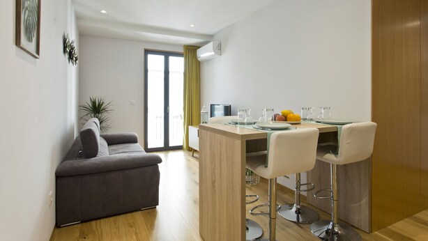 Una sala de estar moderna con un sofá gris, una mesa de comedor de madera con taburetes de bar y un amplio ventanal con cortinas.