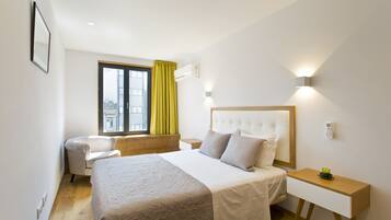 A hotel room with a large bed, a bedside table, a chair, a window with curtains, and a wall-mounted air conditioning unit.