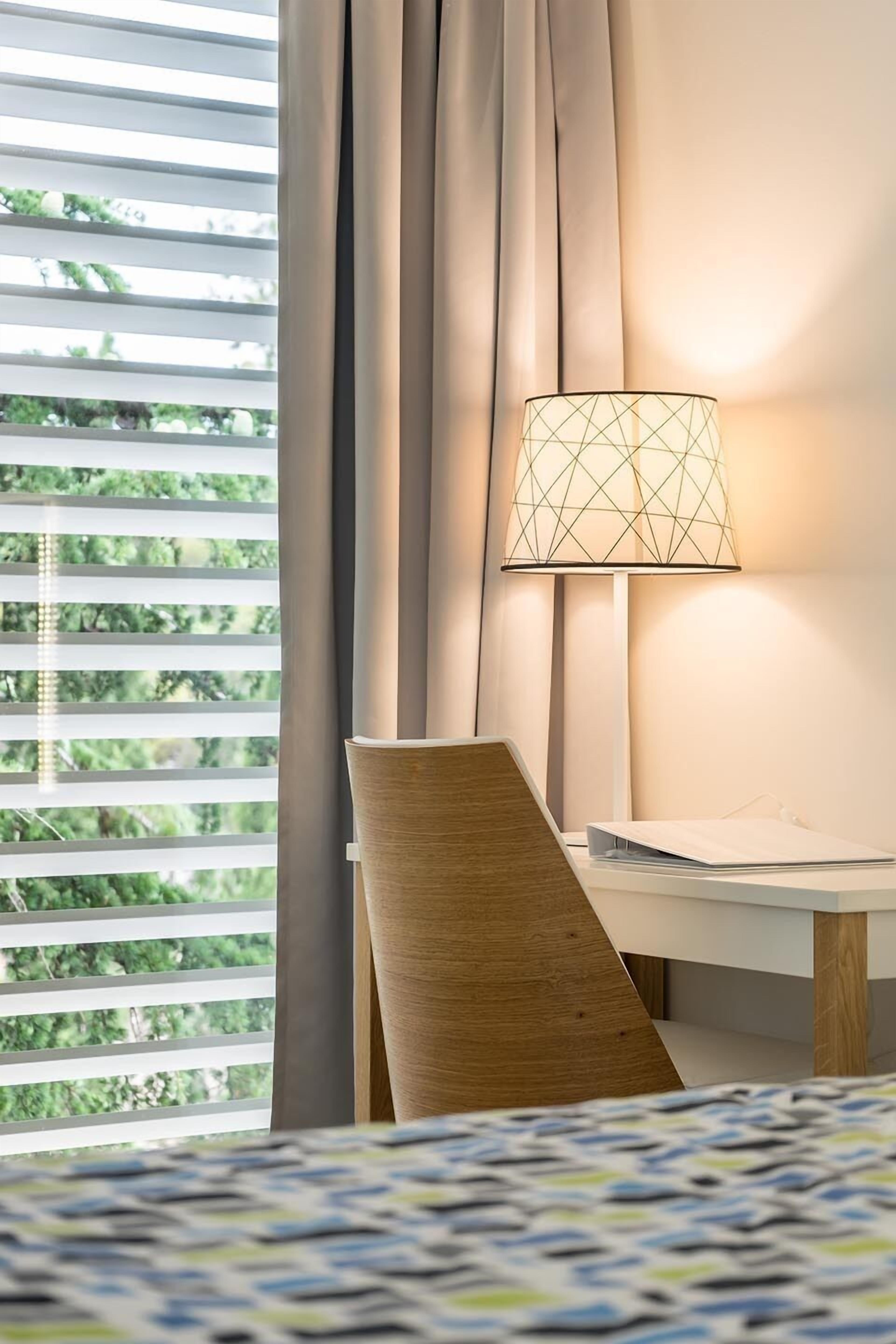 Une chambre d’hôtel avec un lit, un bureau doté d’une lampe et une vue sur la verdure par la fenêtre.