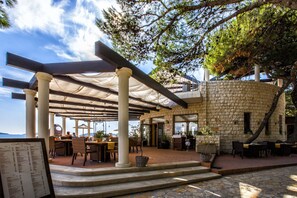 Un área de comedor al aire libre con paredes de piedra, mesas y sillas bajo una pérgola.