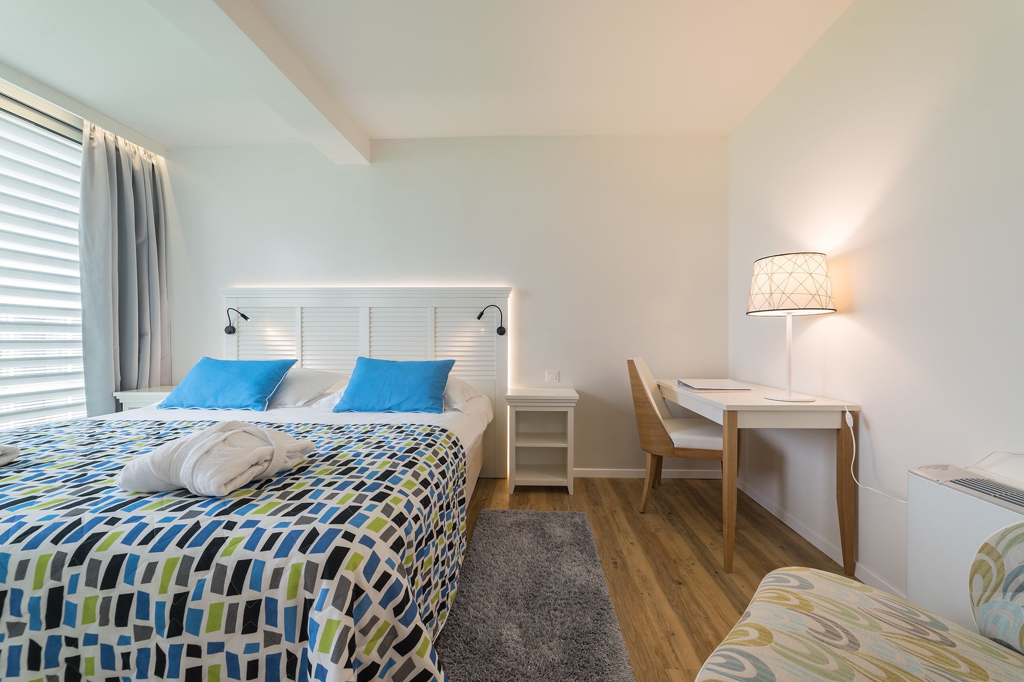 Een slaapkamer met een bed, twee blauwe kussens, een nachtkastje, een bureau, een stoel en een lamp.