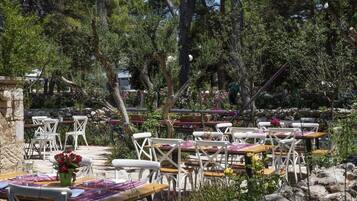 An outdoor dining area with white chairs and tables, surrounded by greenery and trees.