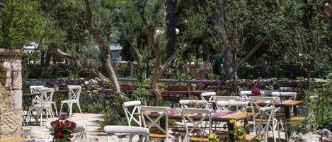 An outdoor dining area with white chairs and tables, surrounded by greenery and trees.