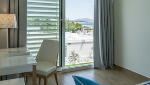 A modern hotel room with a view of a coastal area, a white desk, a chair, and a bed with a blue pillow.