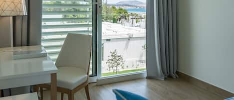A modern hotel room with a view of a coastal area, a white desk, a chair, and a bed with a blue pillow.