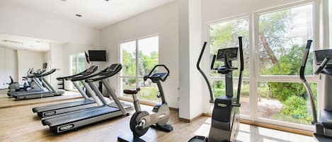 A gym with treadmills, elliptical machines, and a rowing machine, large windows, and a wooden floor.