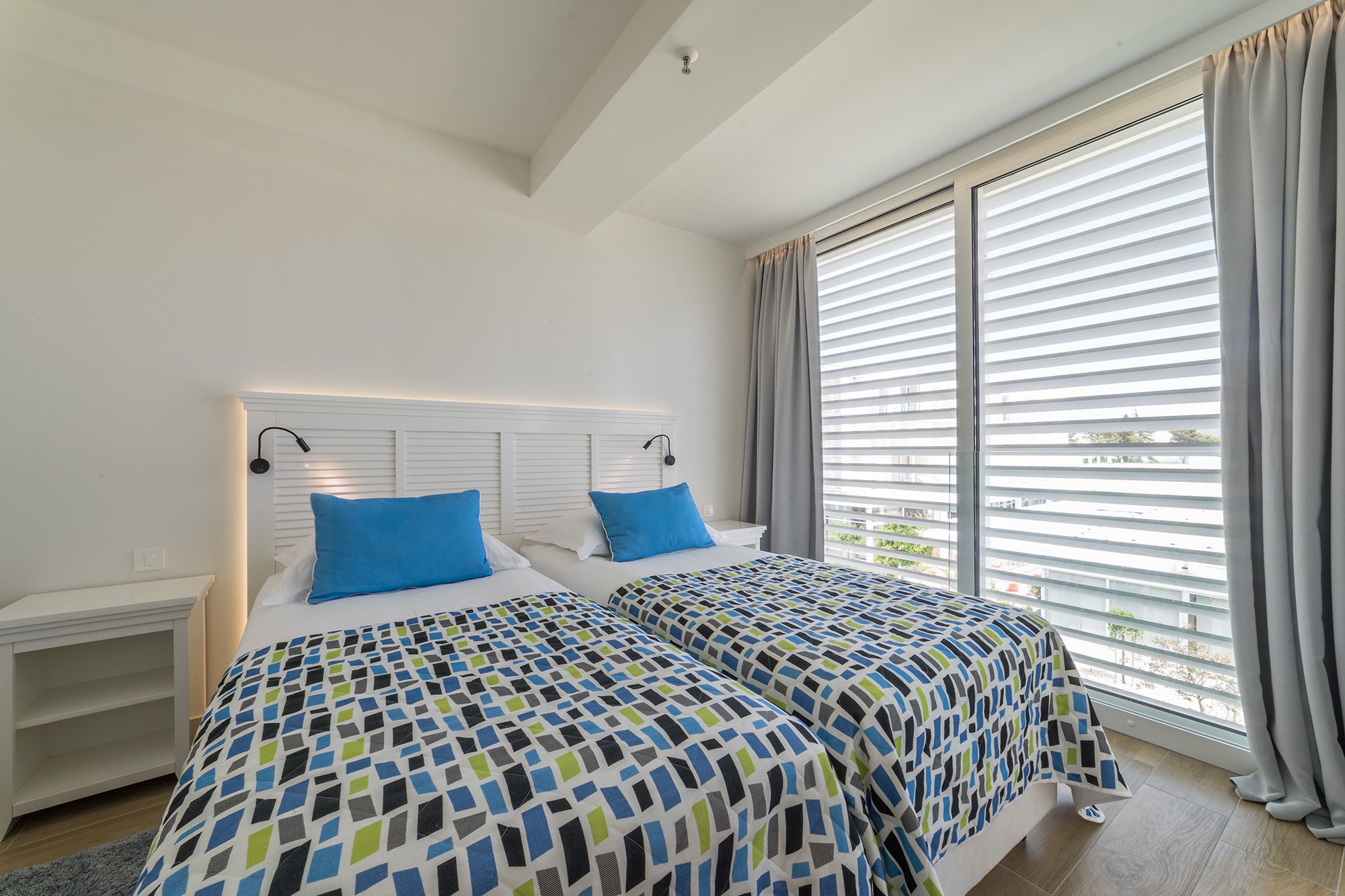 Ein Schlafzimmer mit zwei Betten, weißen Kopfteilen und einem großen Fenster mit Jalousien.