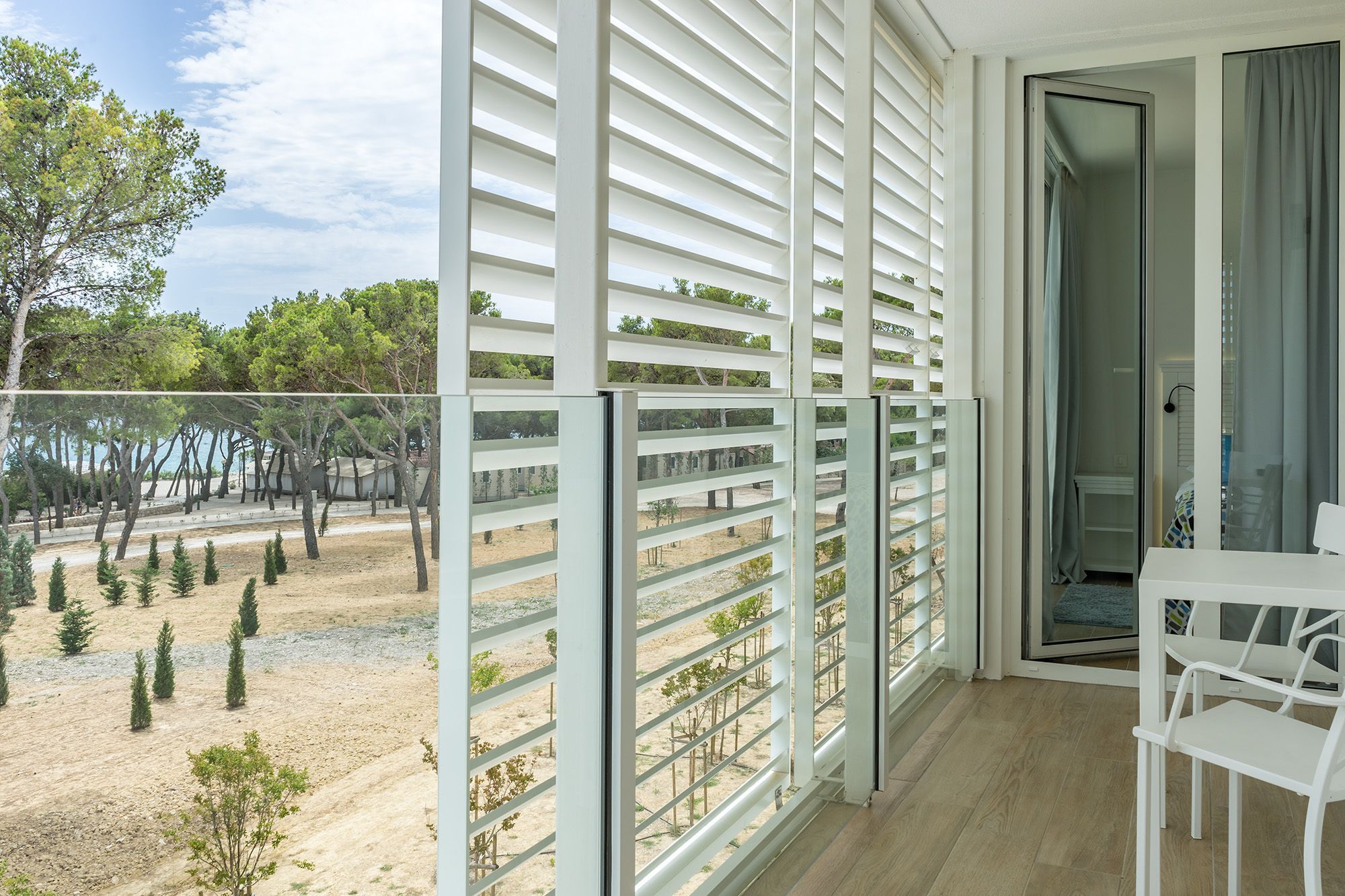 Ein Balkon mit weißen Geländern, einem Tisch mit Stühlen und Blick auf Bäume und einen Strand.