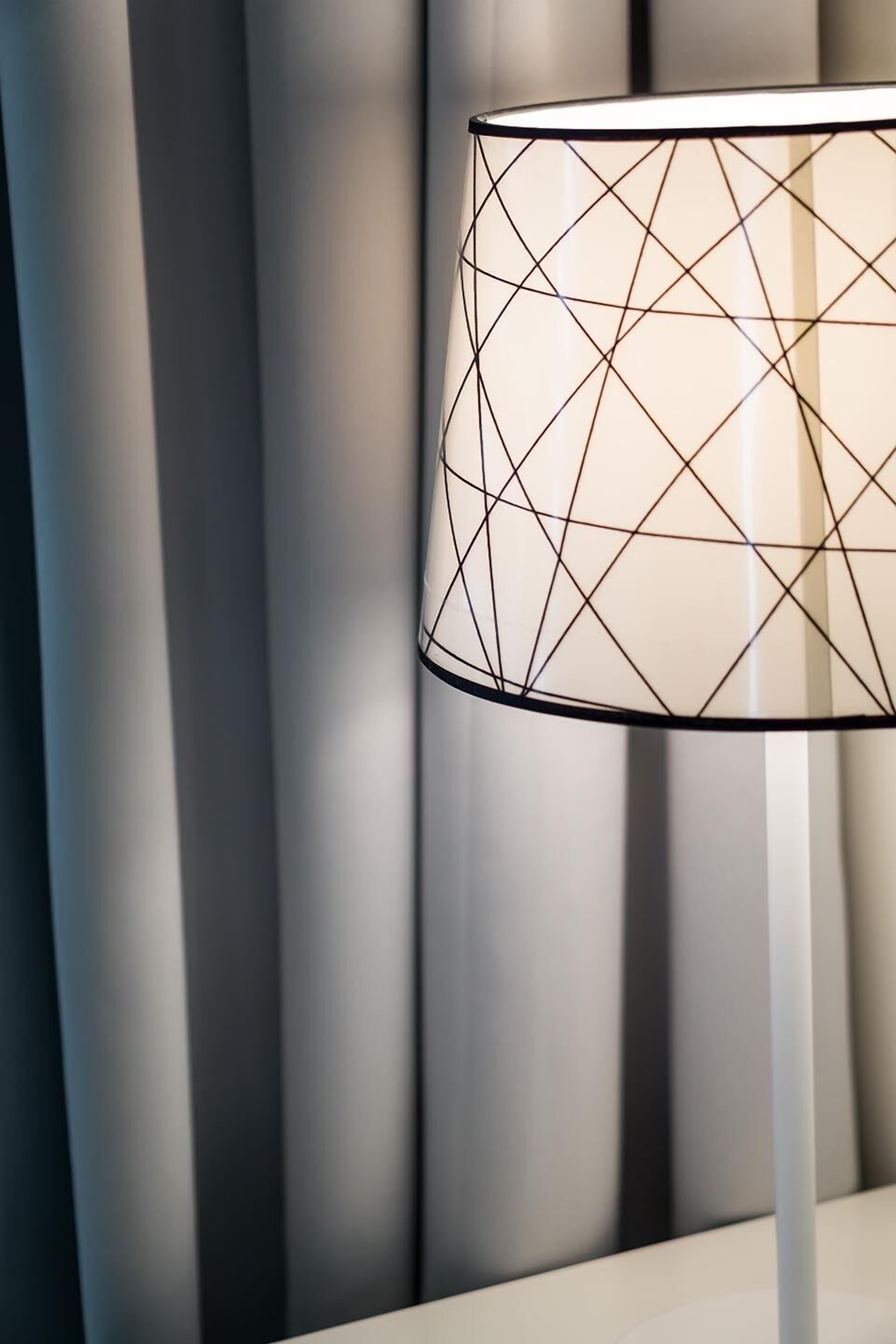A modern lamp with a geometric pattern on its shade, positioned against a backdrop of vertical blinds.