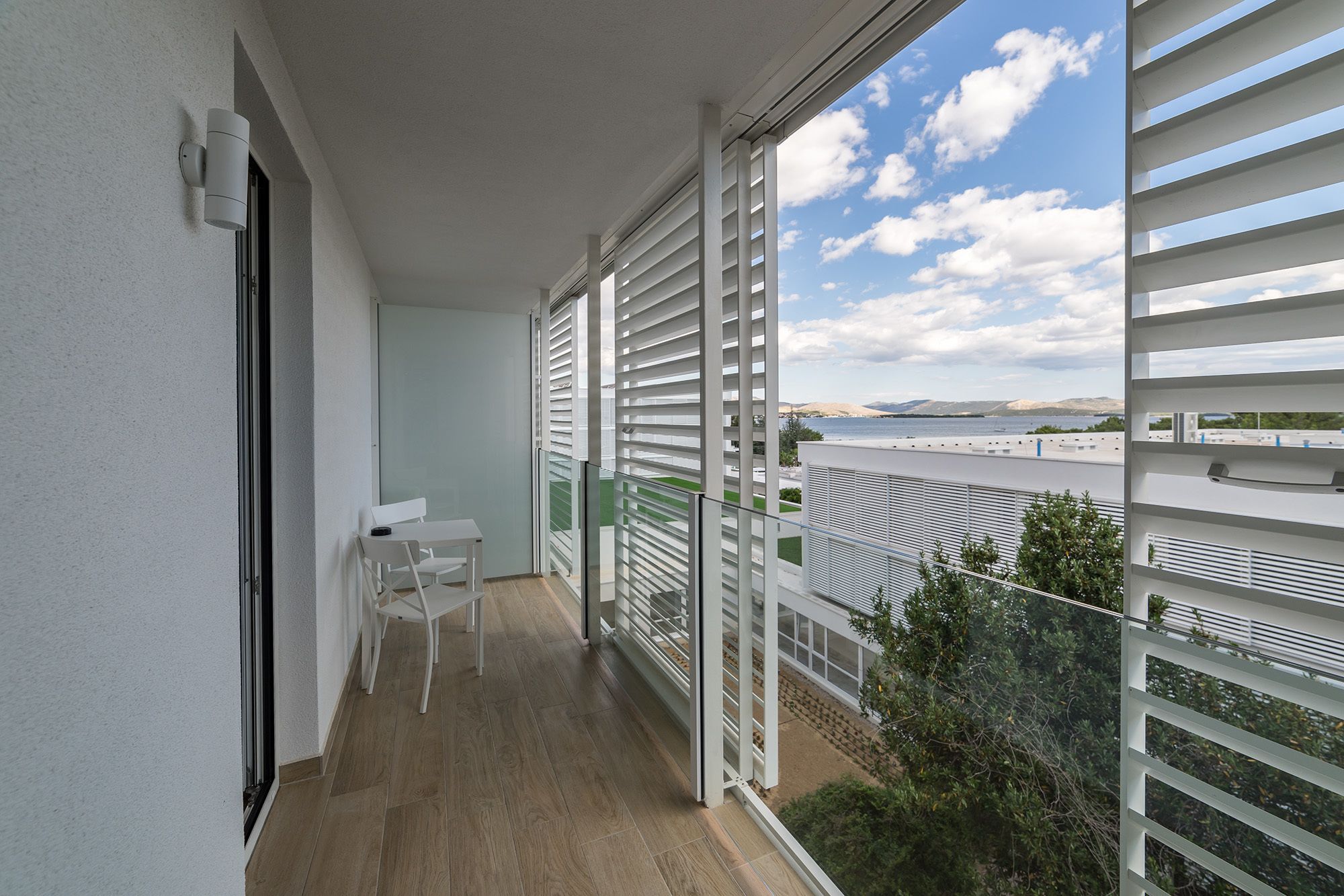 A balcony with a view of a body of water and distant hills, a white table and chair, and a door with a light fixture.