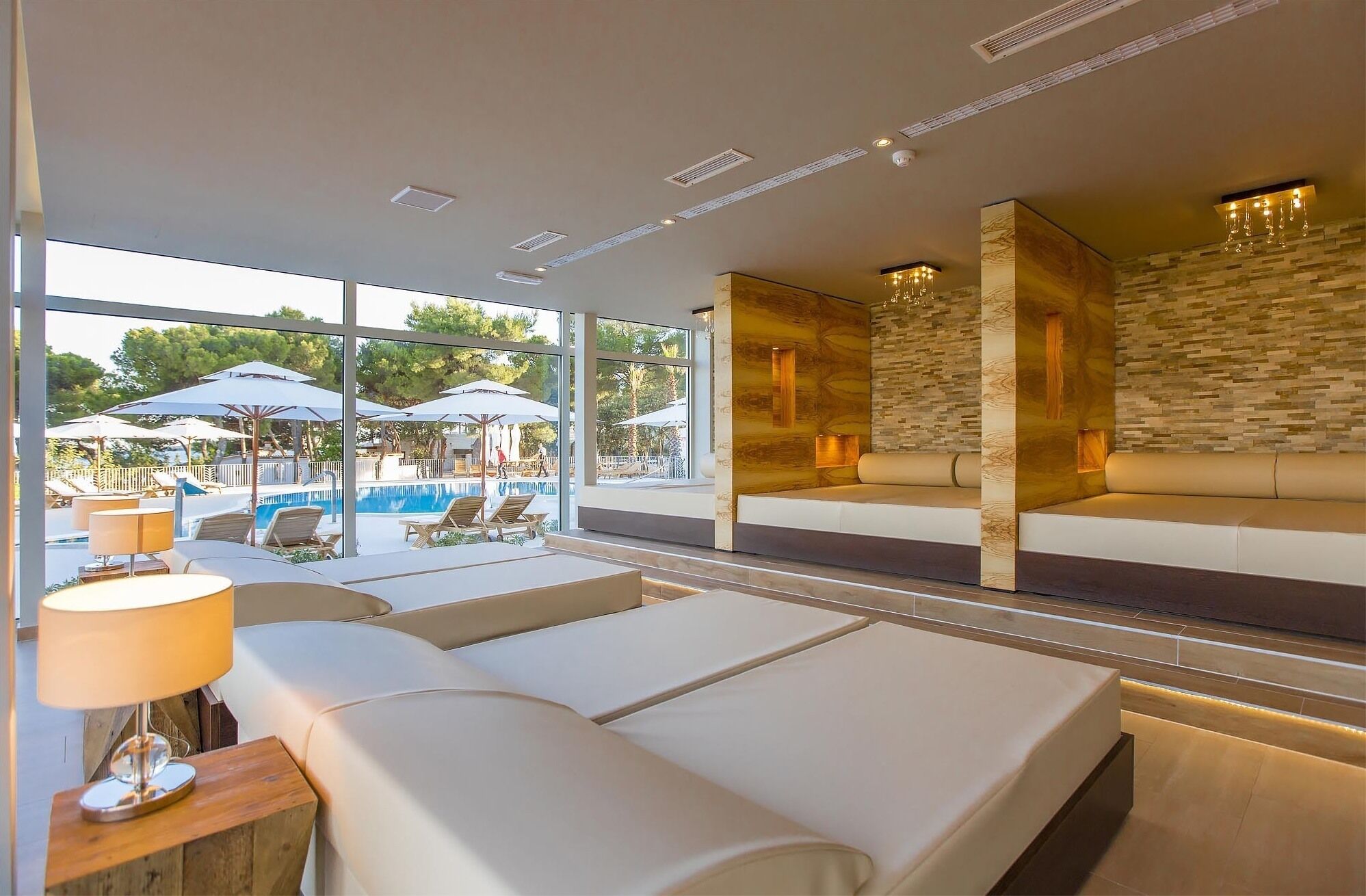 Un área de piscina interior moderna con sillones de descanso y sombrillas, una pared de piedra y un espacio de estar con cojines.