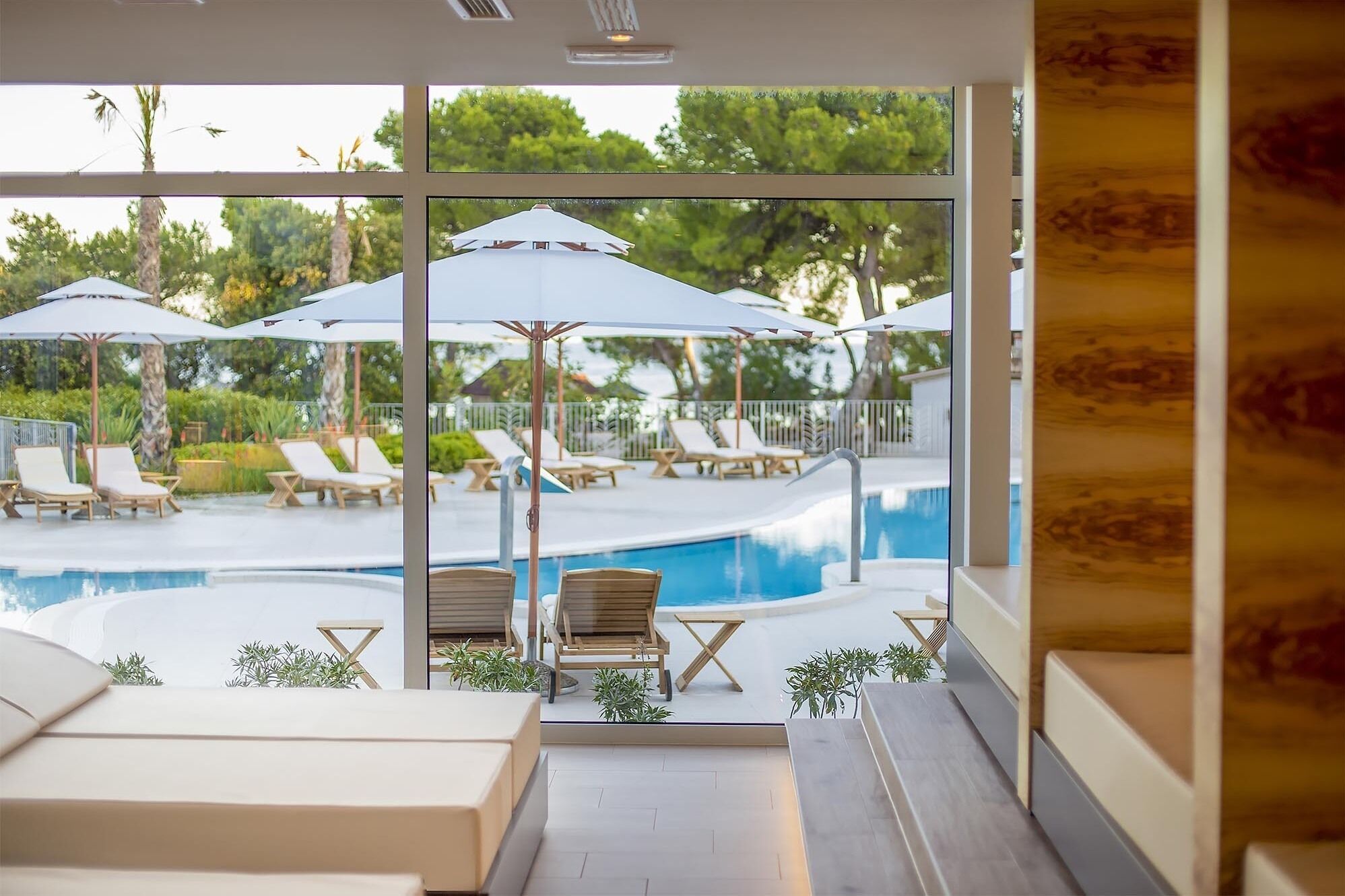 A poolside area with lounge chairs and umbrellas, a swimming pool, and a view of greenery outside.
