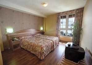 A bedroom with two beds, a bedside table with a lamp, a dresser, and a window with curtains. - Hotel Reino Nevado (Monachil)