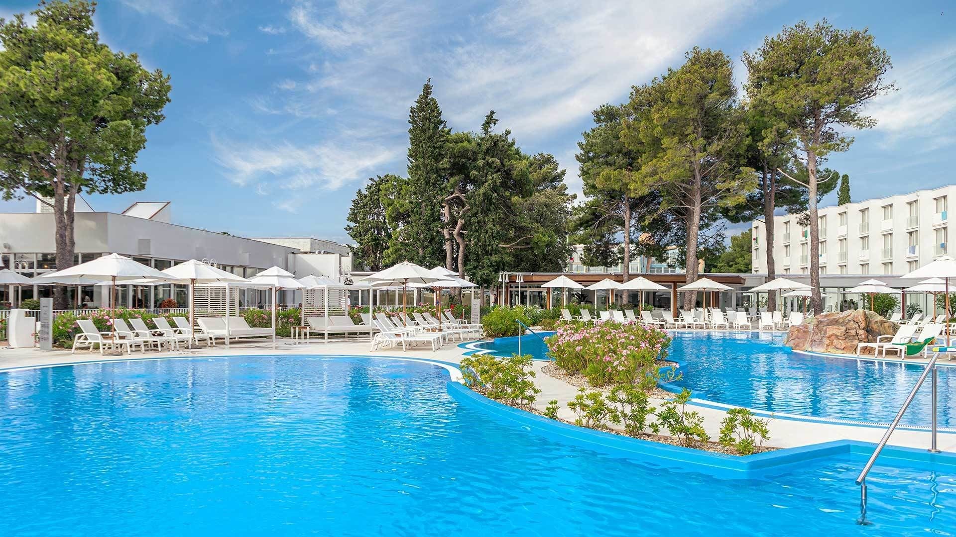 Una piscina con sillones de descanso y sombrillas, rodeada de árboles y un edificio.