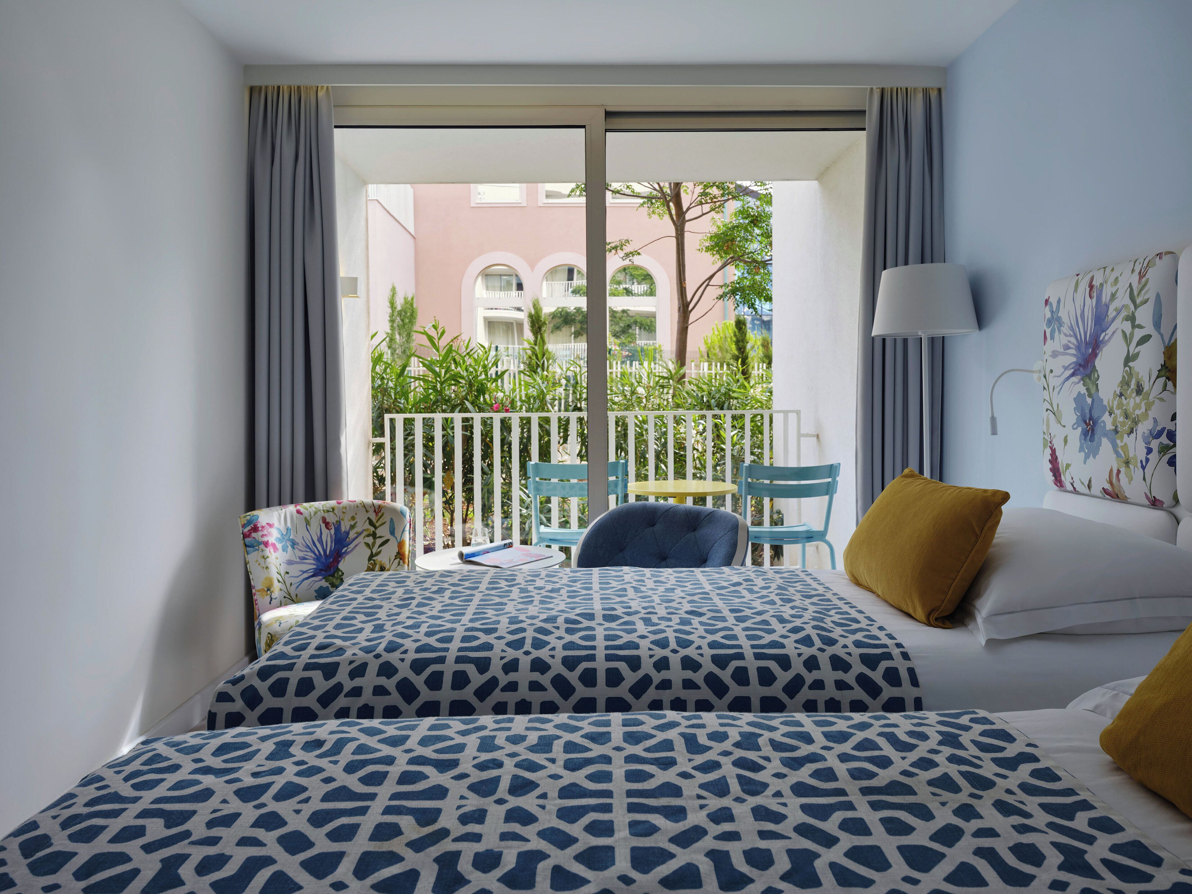 A hotel room with a balcony, a bed with patterned bedding, a chair, a table, and a lamp.