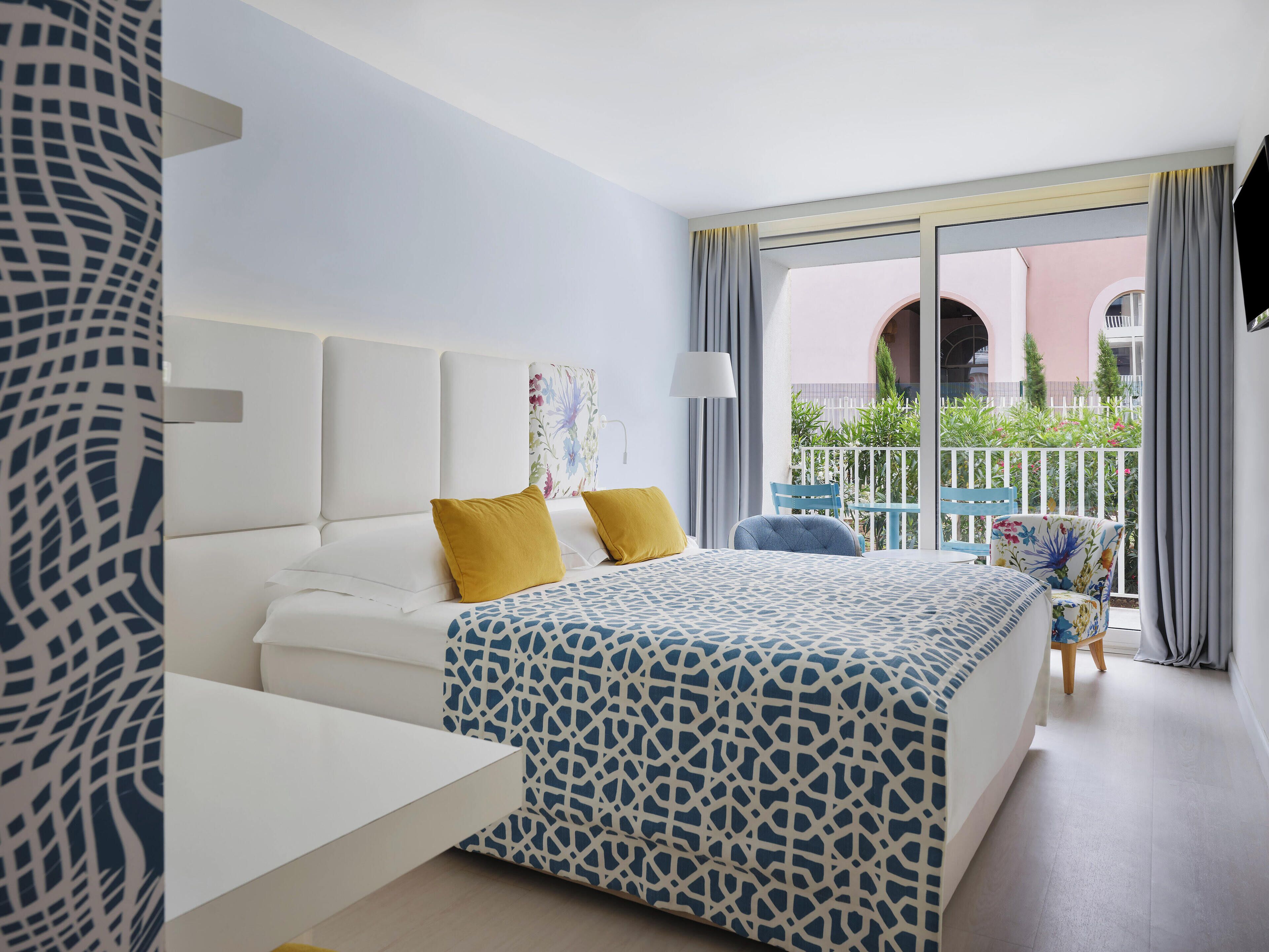 A modern hotel room with a bed, a desk, and a balcony with outdoor seating.