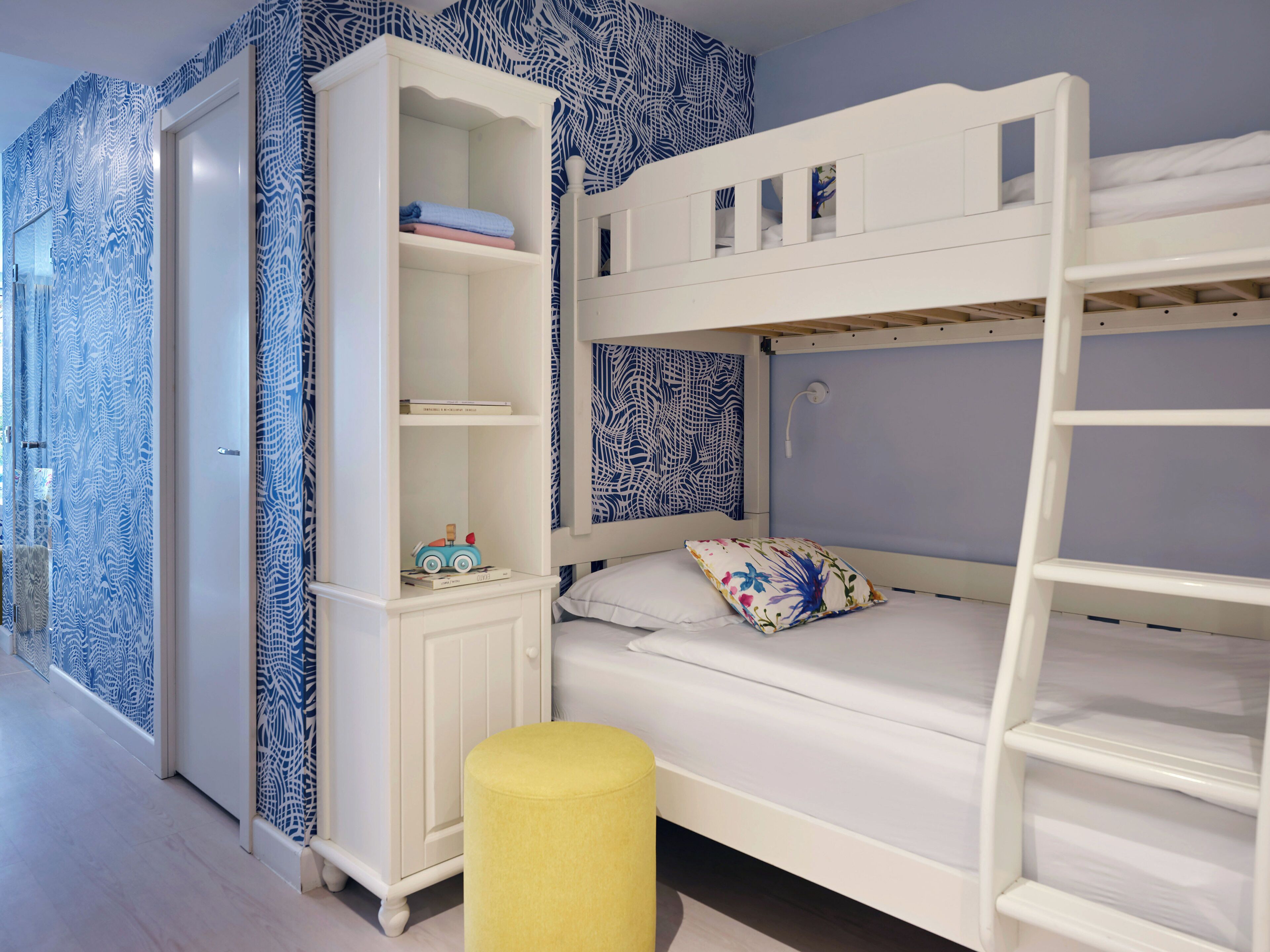 Een stapelbed met een wit hoofdbord, een klein nachtkastje en een gele poef.