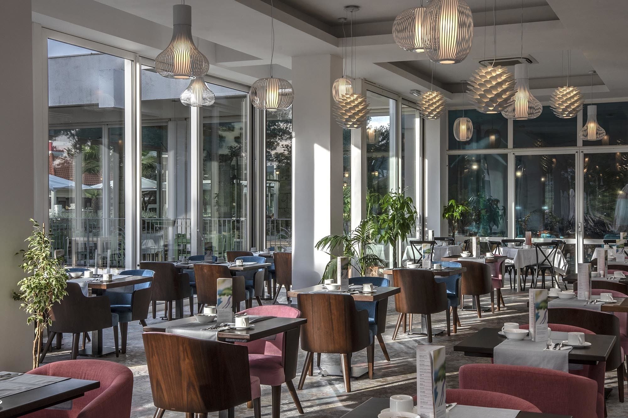 A modern restaurant with large windows, multiple dining tables, and unique hanging light fixtures.