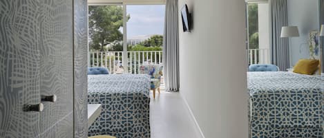 A hotel room with two beds, a balcony with a view, a TV mounted on the wall, and a small table with a lamp.