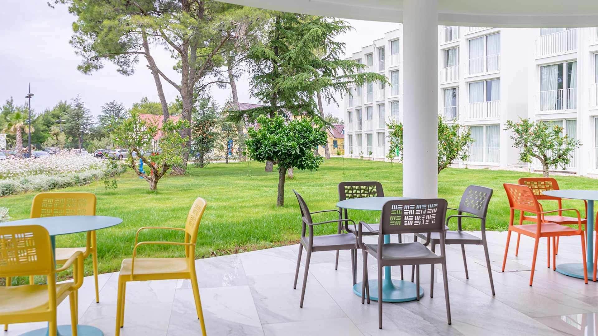 An outdoor patio area with tables and chairs, surrounded by greenery and a building in the background.
