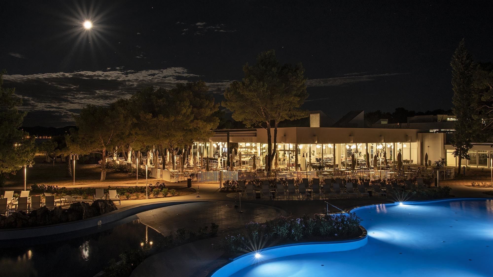 A nighttime view of a resort with a swimming pool, outdoor seating, and a well-lit building with a modern design.