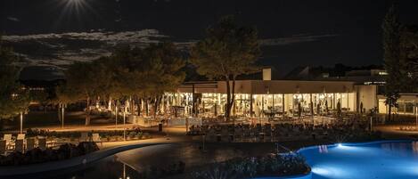 A nighttime view of a resort with a swimming pool, outdoor seating, and a well-lit building with a modern design.