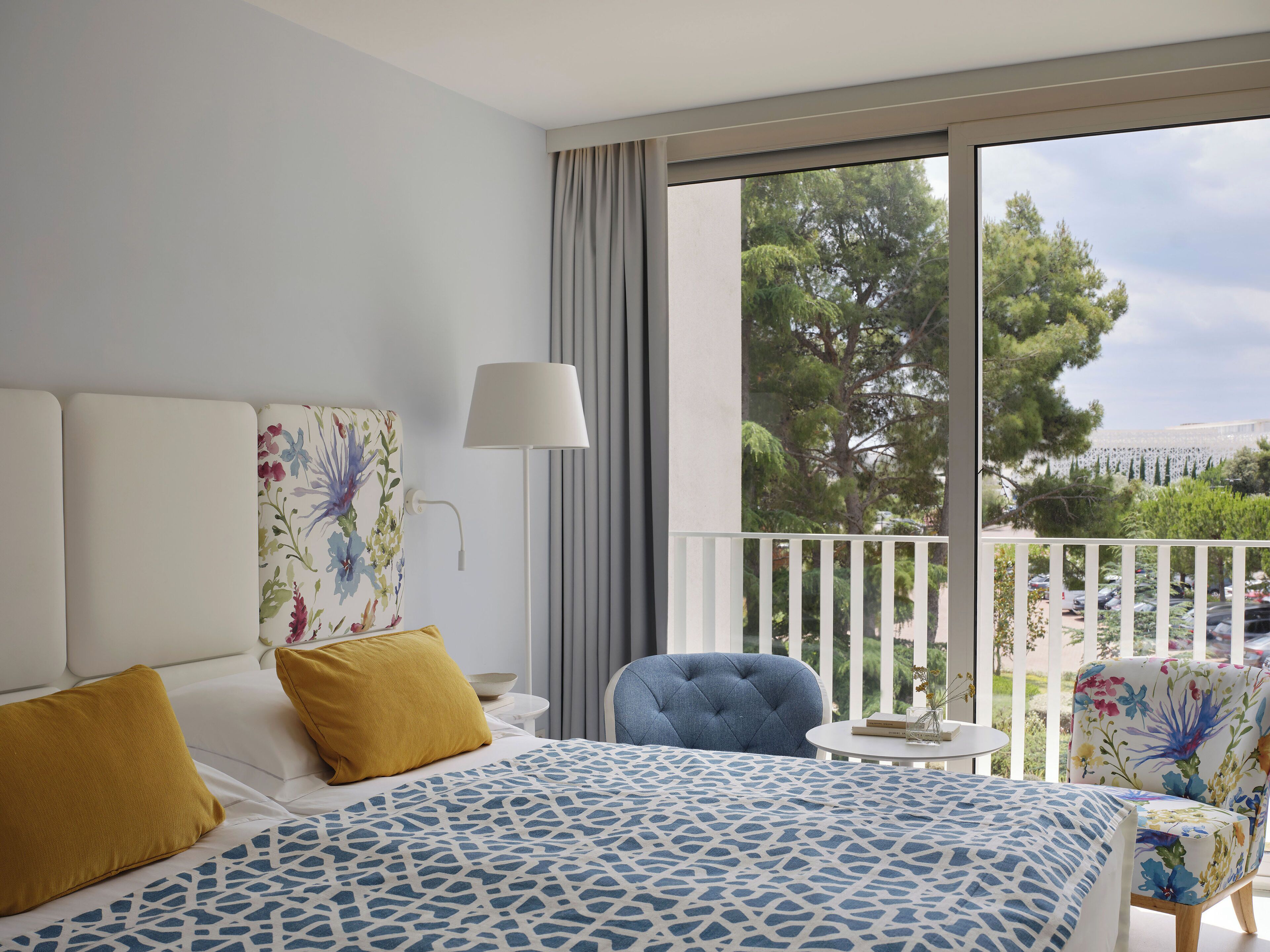 Habitación de hotel con cama, sillón, mesa y balcón con vista a árboles y coches aparcados.