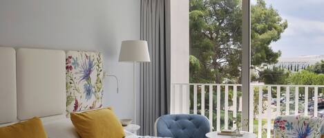 Habitación de hotel con cama, sillón, mesa y balcón con vista a árboles y coches aparcados.