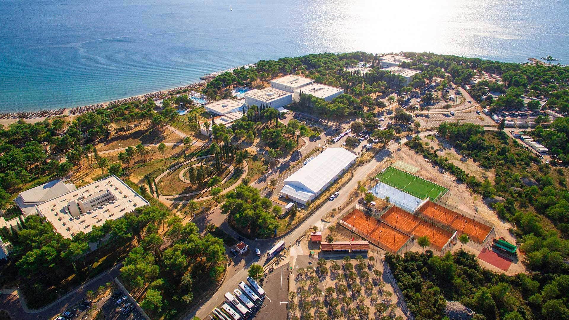 An aerial view of a coastal area with a large building complex, a beach, and a sports field.